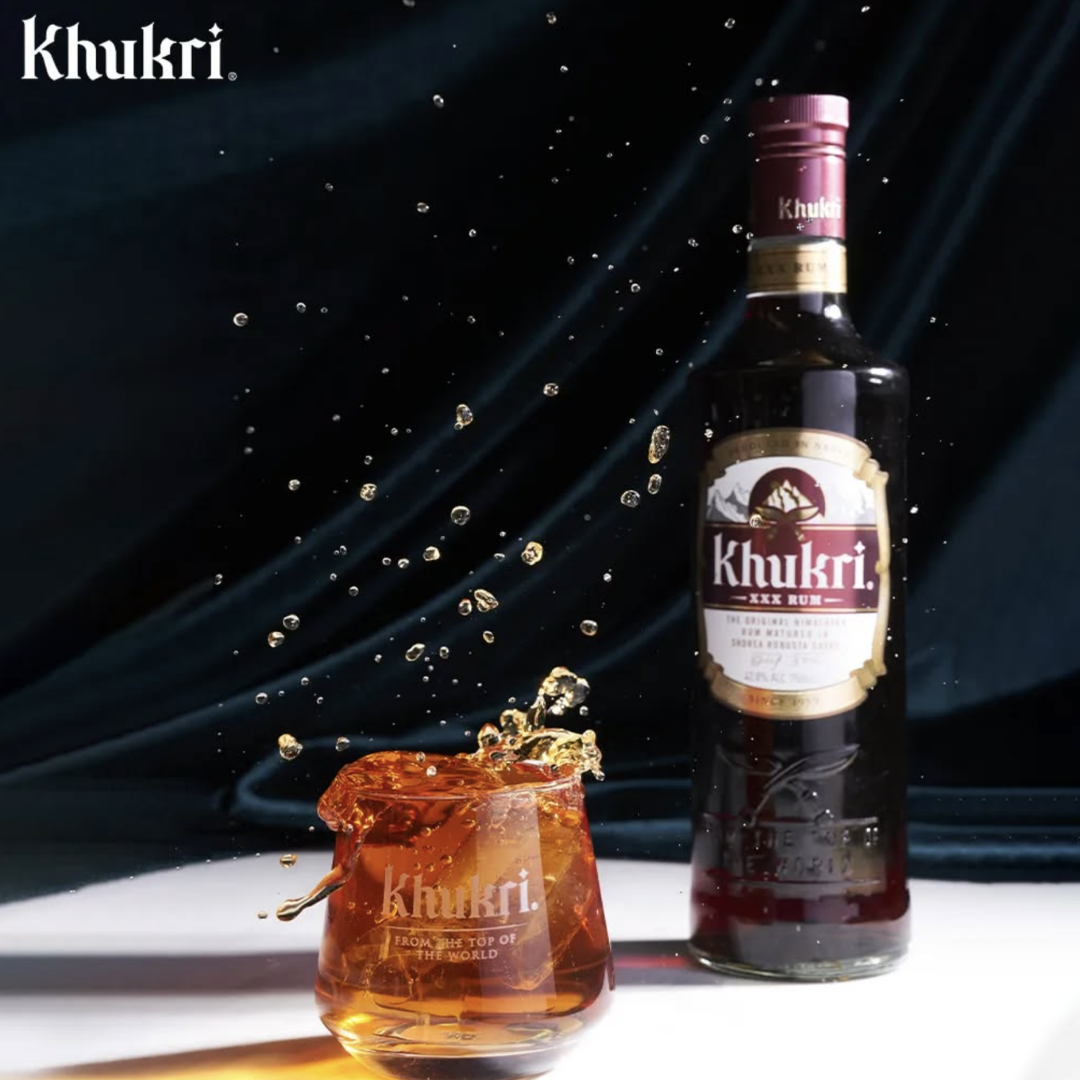 A premium Khukuri Rum bottle placed beside a glass filled with dark rum and ice, captured mid-splash against a dark elegant background. The image highlights the rich amber color of Khukuri XXX Rum, showcasing its bold character and premium Nepali craftsmanship, ideal for representing Khukuri rum price in Nepal and authentic local rum quality.